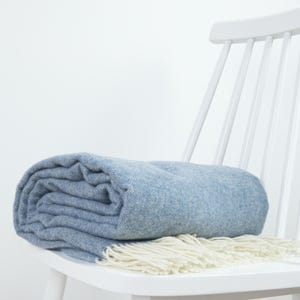 May include: A folded light blue throw blanket with a woven herringbone pattern and cream-coloured fringe along the edge. The blanket is draped over the seat of a white wooden chair against a plain white background.