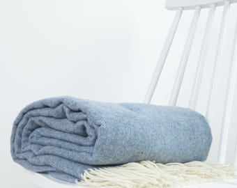Puede incluir: Una manta de color azul claro doblada con un patrón de espiga tejido y flecos de color crema a lo largo del borde. La manta está colocada sobre el asiento de una silla de madera blanca contra un fondo blanco liso.