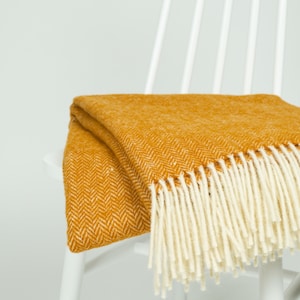 May include: Mustard yellow throw blanket with a herringbone pattern and cream-colored fringe, draped over a white chair. The blanket's texture and color create a cozy, inviting aesthetic. A decorative home textile.