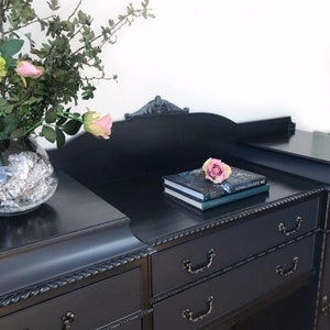 May include: An antique dresser painted black with intricate details and carved trim. The dresser has six drawers with black metal handles. The dresser is sitting on a wooden floor.
