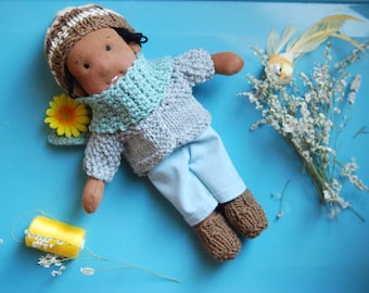 May include: A handmade doll with brown skin, wearing a grey knitted sweater, a light blue scarf, and light blue pants. The doll is wearing brown knitted boots and has a white and brown striped hat.