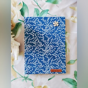 May include: A blue journal with a white leaf pattern and a wooden flower closure. The journal has a brown leather tag with the word "Handmade" on it. The journal is on a floral patterned surface.