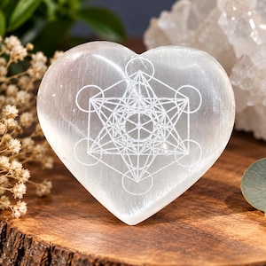 Op de afbeelding: Een hartvormig stuk doorschijnend wit selenietkristal met een fijn gegraveerd geometrisch Metatron-kubuspatroon op het vooroppervlak. De steen heeft een gladde, gepolijste afwerking en een helder, kristallijn uiterlijk.