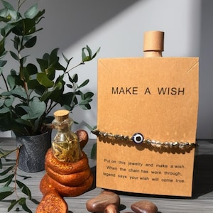 May include: A brown card with "MAKE A WISH" printed on it, a beaded bracelet with an evil eye charm, and a small glass bottle with a cork stopper. The image also includes a decorative snake and some wooden stones.