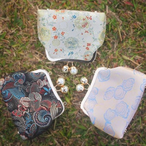 Puede incluir: Tres pequeñas carteras de tela con cierres dorados y detalles de perlas. Las carteras están decoradas con motivos florales en tonos azules, blancos y negros.