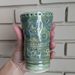 May include: A green ceramic mug with a floral design. The mug has a textured surface and a slightly flared rim.