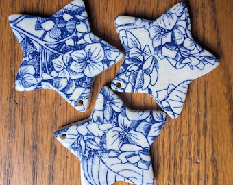 Op de afbeelding: Drie stervormige keramische ornamenten met een blauw bloemmotief op een witte achtergrond. Elk ornament heeft een klein gaatje om op te hangen. De ornamenten zijn gerangschikt op een houten oppervlak.