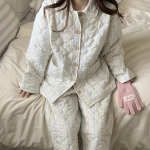 May include: A quilted pajama set in a floral pattern with light blue stripes. The set includes a button-down jacket with pockets and matching pants. A pink glove rests on the bed. The pajamas are made of a soft, comfortable material.