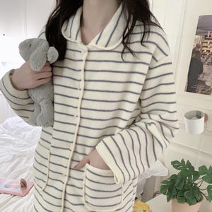 Peut inclure: Ensemble de pyjama crème à rayures horizontales grises. Le haut à manches longues a un col, une fermeture boutonnée et deux poches avant. Le pantalon assorti présente également le même motif rayé. La personne tient un animal en peluche gris.
