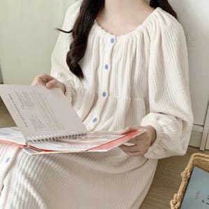 Peut inclure: Robe crème à manches longues avec un devant boutonné et un col rond. La robe a un aspect texturé, semblable à du velours côtelé, et des boutons bleu clair. La personne tient un livre à reliure spirale. Un panier en osier avec un livre est en arrière-plan.