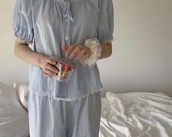 Puede incluir: Conjunto de pijama azul claro con estampado de lunares. La parte superior tiene mangas cortas abullonadas y un escote redondo con ribete de encaje. Los pantalones a juego son de pernera ancha. Una goma de pelo blanca está en la muñeca.