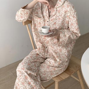 Könnte beinhalten: Cremefarbenes Blumen-Pyjama-Set mit einem Muster aus kleinen rosa Blüten. Das Set besteht aus einem Hemd mit Knöpfen und einer Tasche sowie einer passenden Hose. Eine kleine weiße Tasse und Untertasse werden gehalten. Der Pyjama ist aus einem weichen, leichten Stoff.