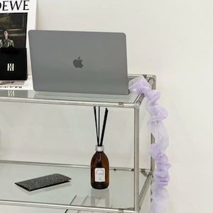 Può includere: Un'unità a scaffale in metallo argentato con ripiani in vetro. Un computer portatile, una piccola bottiglia di olio per diffusore con canne nere, un taccuino bianco e nero e una pila di libri sono sugli scaffali. Una ghirlanda di tulle color lavanda è appesa sul lato dell'unità a scaffale.