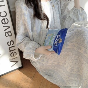 May include: A light blue floral patterned dress with long sleeves. The dress has a round neckline and a button closure. A bag of Scotti snack chips is held in the hand. The text "Viens avec" is visible on a sign.
