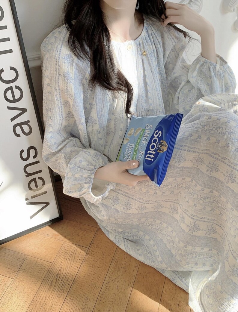May include: A light blue floral patterned dress with long sleeves. The dress has a round neckline and a button closure. A bag of Scotti snack chips is held in the hand. The text "Viens avec" is visible on a sign.