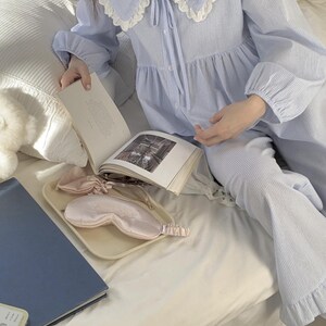 May include: A light blue striped pajama set with a white lace collar and a bow tie. The set includes a long-sleeved top and matching pants. The pajamas are laid out on a white bed with a book and a sleep mask.