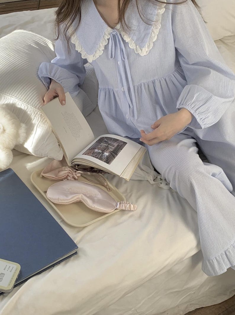 May include: A light blue striped pajama set with a white lace collar and a bow tie. The set includes a long-sleeved top and matching pants. The pajamas are laid out on a white bed with a book and a sleep mask.