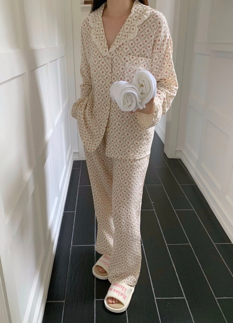 May include: A white and pink floral print pajama set with a collared top and long pants. The top has a frilled collar and button closure. The pants have a wide leg style. The model is wearing white slippers with the text "Relax Home" printed on them.
