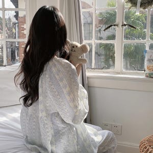 May include: A person wearing a light blue and white patterned pajama set, holding a small teddy bear. The pajamas have a floral design. The person is sitting on a white bed near a window with a view of a building.