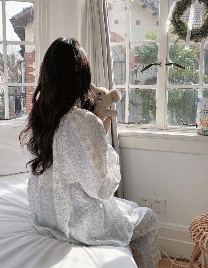 May include: A person wearing a light blue and white patterned pajama set, holding a small teddy bear. The pajamas have a floral design. The person is sitting on a white bed near a window with a view of a building.