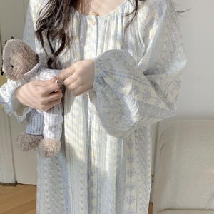 May include: A pale blue and white floral print nightdress with long sleeves and a button-down front. The nightdress is being held. A small teddy bear in a striped shirt is also in the image.