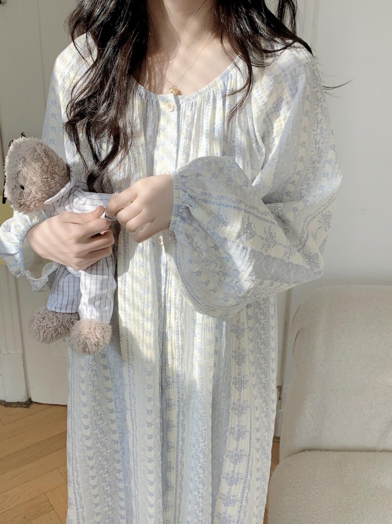 May include: A pale blue and white floral print nightdress with long sleeves and a button-down front. The nightdress is being held. A small teddy bear in a striped shirt is also in the image.