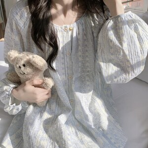 May include: A light blue and white floral patterned pajama set. The top has a button-down front, round neckline, and long, puffy sleeves. The matching trousers are visible. A small, beige teddy bear is held in the hand.