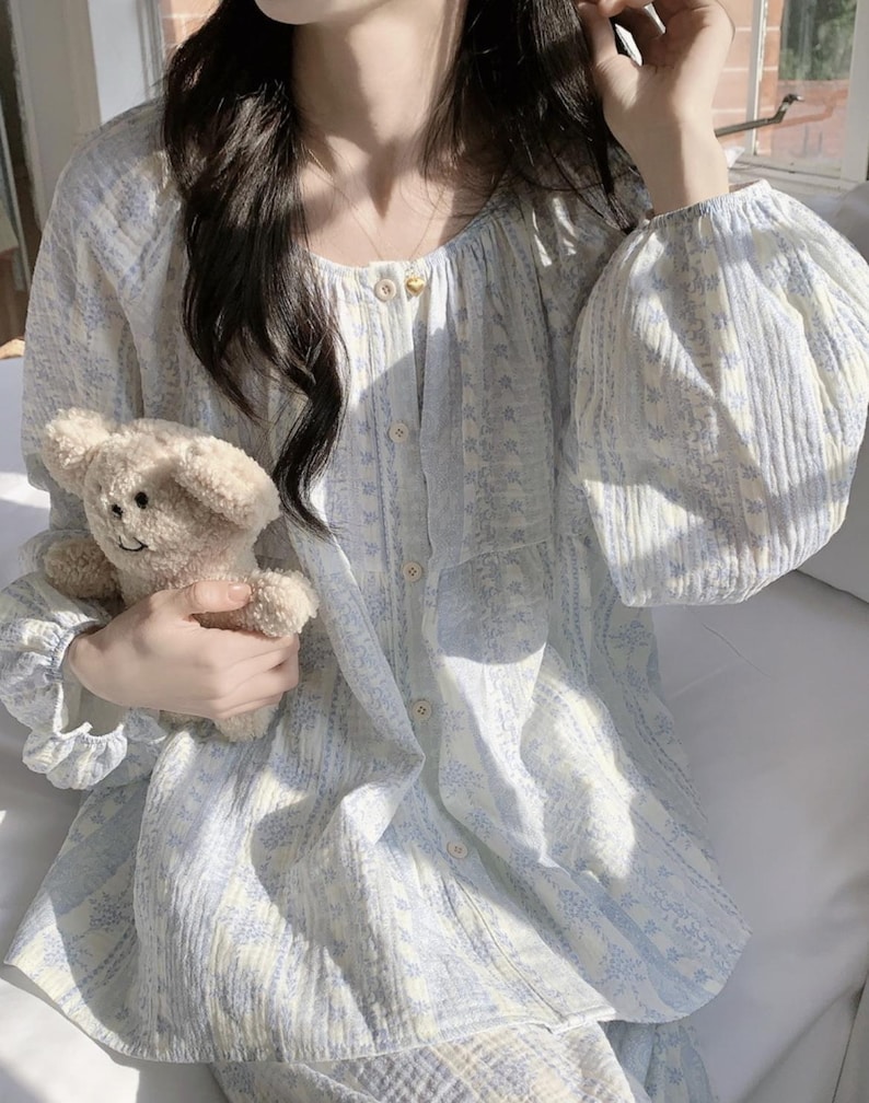 May include: A light blue and white floral patterned pajama set. The top has a button-down front, round neckline, and long, puffy sleeves. The matching trousers are visible. A small, beige teddy bear is held in the hand.