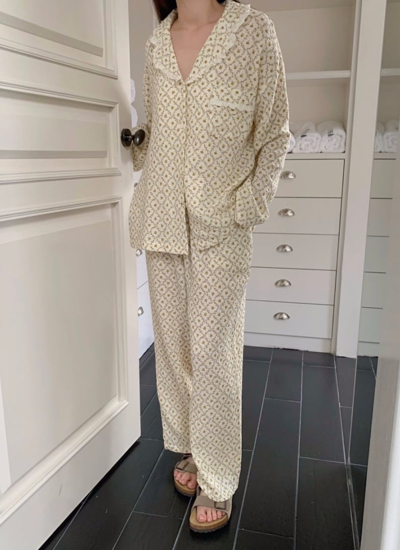 May include: A white pajama set with a floral pattern. The top has a lace trim collar and long sleeves. The pants are long and have a straight leg.
