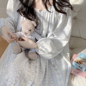 May include: A pale blue floral print nightdress with long sleeves and a button-down front. The nightdress is held with a small teddy bear in a striped shirt. A packet of biscuits and a pink book are also visible.