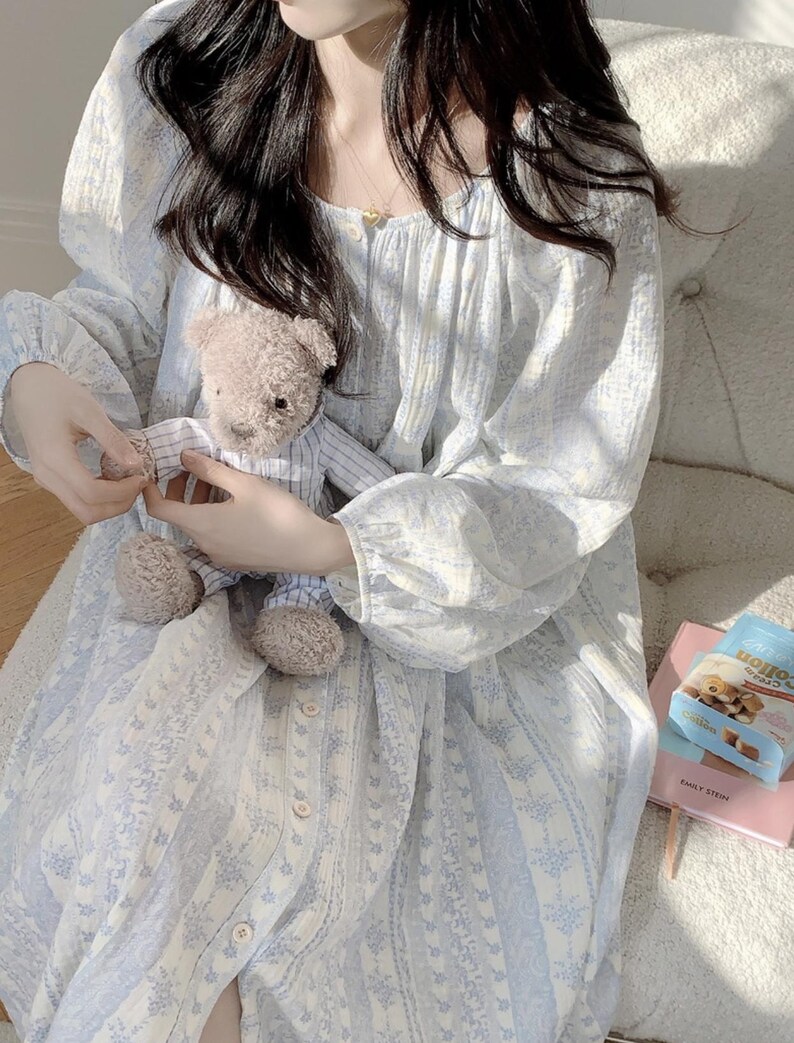 May include: A pale blue floral print nightdress with long sleeves and a button-down front. The nightdress is held with a small teddy bear in a striped shirt. A packet of biscuits and a pink book are also visible.