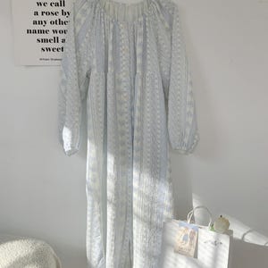 May include: A light blue and white patterned dress with long sleeves hangs on a hanger. The dress has a round neckline and buttons down the front. A white shopping bag with a flower sits below the dress. A poster with text from Romeo and Juliet is on the wall.