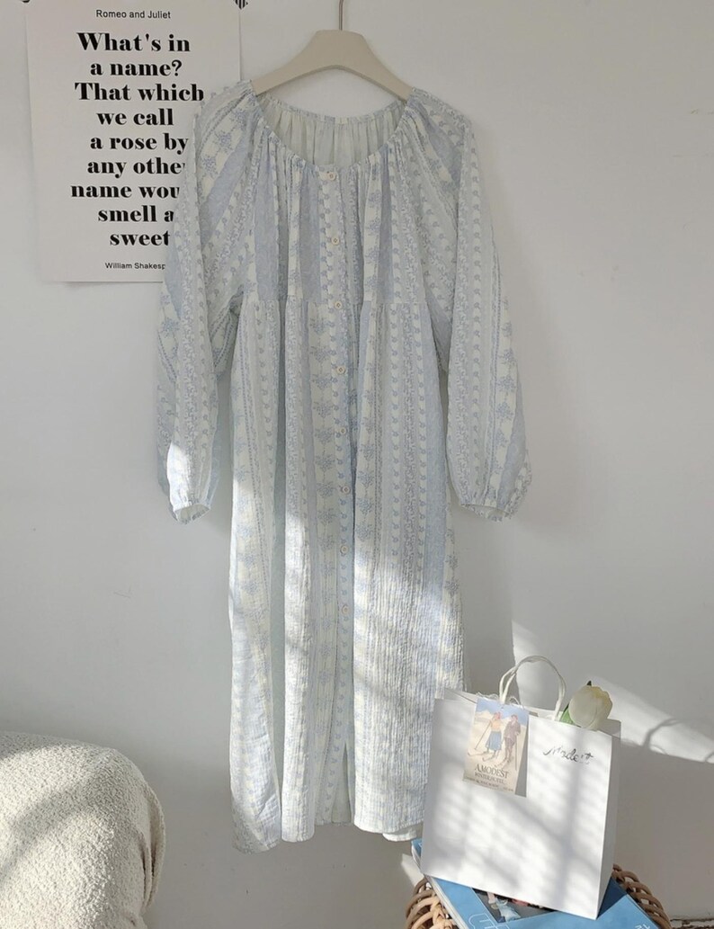 May include: A light blue and white patterned dress with long sleeves hangs on a hanger. The dress has a round neckline and buttons down the front. A white shopping bag with a flower sits below the dress. A poster with text from Romeo and Juliet is on the wall.