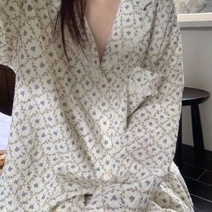 May include: A white long-sleeved shirt with a floral pattern and a lace collar. The shirt has a button-up front and a pocket on the left side.
