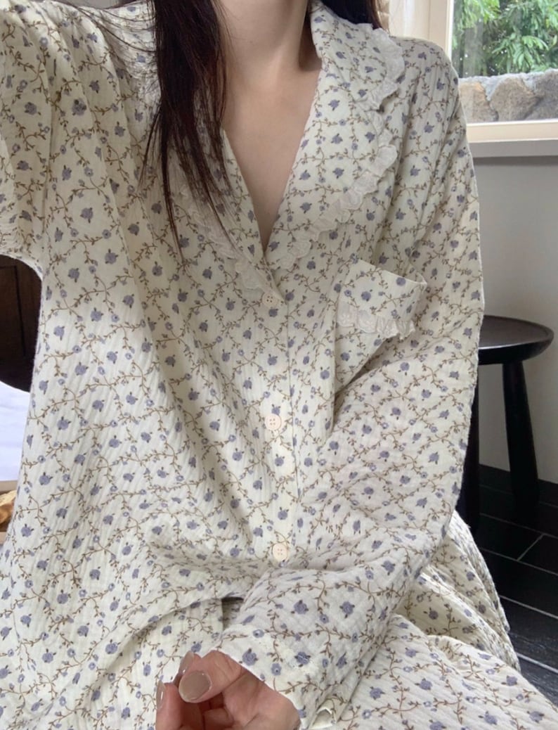May include: A white long-sleeved shirt with a floral pattern and a lace collar. The shirt has a button-up front and a pocket on the left side.