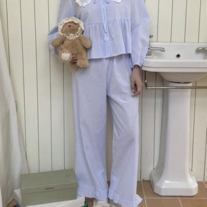 May include: A light blue pajama set with a white lace collar and a ruffled hem. The top has a button-up front and a bow tie. The pants are wide-legged and have a striped pattern.