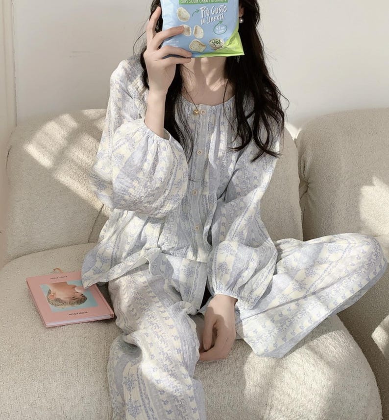 May include: A light blue and white floral patterned pajama set. The top has long sleeves and a button-down front, and the trousers have a relaxed fit. A small pink book and a bag of crisps are also visible.
