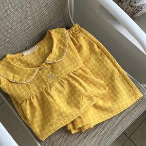 Könnte beinhalten: Gelbes Pyjama-Set mit strukturiertem Muster. Das Oberteil hat einen weiß eingefassten Peter Pan-Kragen und einen Knopfverschluss. Die passende Hose ist ordentlich neben dem Oberteil gefaltet. Das Set ist aus weichem, leichtem Stoff.