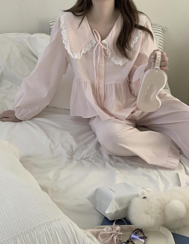 May include: A pink pajama set with a white lace collar and a bow tie. The set includes a long-sleeved top and matching pants. The model is holding a white eye mask.