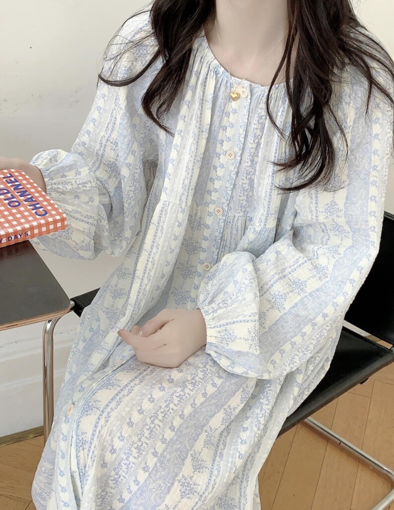 May include: A light blue and white floral patterned dress with long sleeves and button details. The dress has a round neckline and a relaxed fit. The sleeves are gathered at the cuffs. The dress is made of a lightweight fabric.