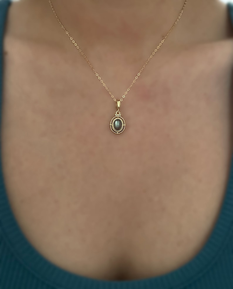 May include: A gold chain necklace with a pendant featuring a grey oval gemstone surrounded by a gold beaded border.