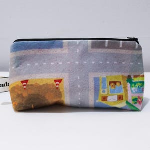 Puede incluir: Una bolsa rectangular con cremallera y un estampado colorido que representa una escena de carretera con edificios, vehículos y conos de tráfico. La bolsa tiene una cremallera negra y una pequeña etiqueta blanca con texto negro.