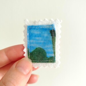 May include: A miniature fabric postage stamp with a landscape scene. The stamp features a blue sky and green hills, framed by a white, scalloped edge. The image is hand-stitched with a light blue thread.
