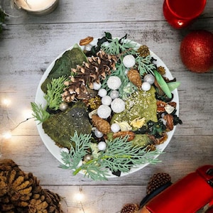 May include: A white plate displays a Christmas arrangement of pinecones, green foliage, white baubles, and silver decorations. The display is on a light wood surface, with a candle and red baubles nearby, creating a festive scene.