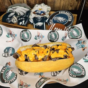 May include: A yellow bread roll filled with meat and topped with melted yellow cheese, presented on a paper with Philadelphia Eagles logos. In the background are Philadelphia Eagles themed cookies and decorations.