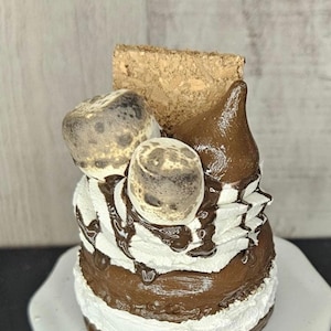 May include: A decorative dessert featuring layers of white frosting and chocolate, topped with toasted marshmallows, chocolate drizzle, and a piece of graham cracker. The dessert sits on a white plate.