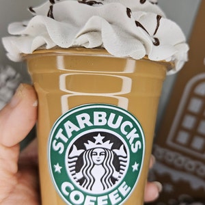 Puede incluir: Un vaso de caf&eacute; Starbucks marr&oacute;n y beige con crema batida y un chorrito de chocolate en la parte superior. El vaso tiene el logotipo de Starbucks con el texto "Starbucks Coffee".