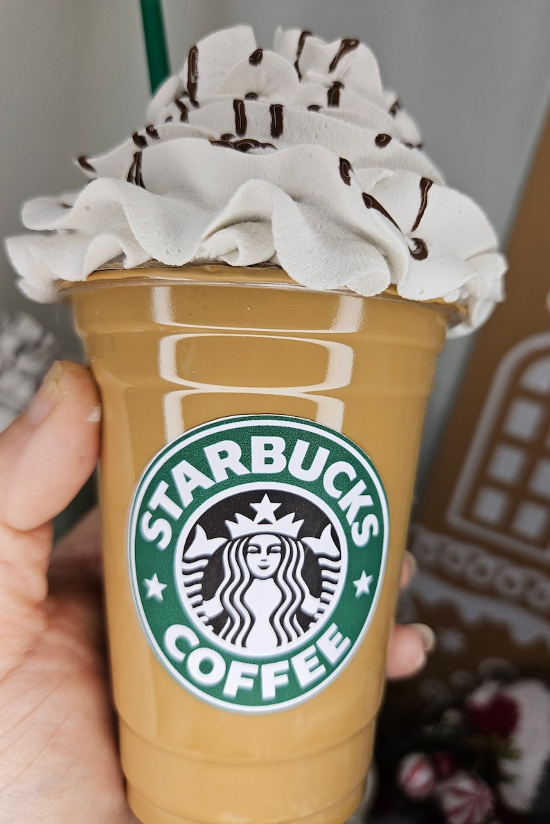 Puede incluir: Un vaso de caf&eacute; Starbucks marr&oacute;n y beige con crema batida y un chorrito de chocolate en la parte superior. El vaso tiene el logotipo de Starbucks con el texto "Starbucks Coffee".
