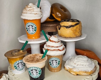 May include: A tiered display with fall-themed treats, including iced coffee, cinnamon rolls, and pumpkin spice lattes. The coffee cups feature the Starbucks logo and the text "Starbucks Coffee".