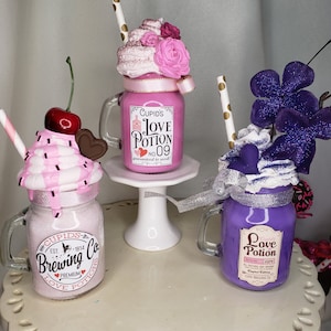 May include: Three decorative mason jar-shaped containers with whipped cream toppings, straws, and labels reading "Love Potion." One is pink with a cherry, another is purple, and the third is pink with the text "Cupid's Brewing Co."